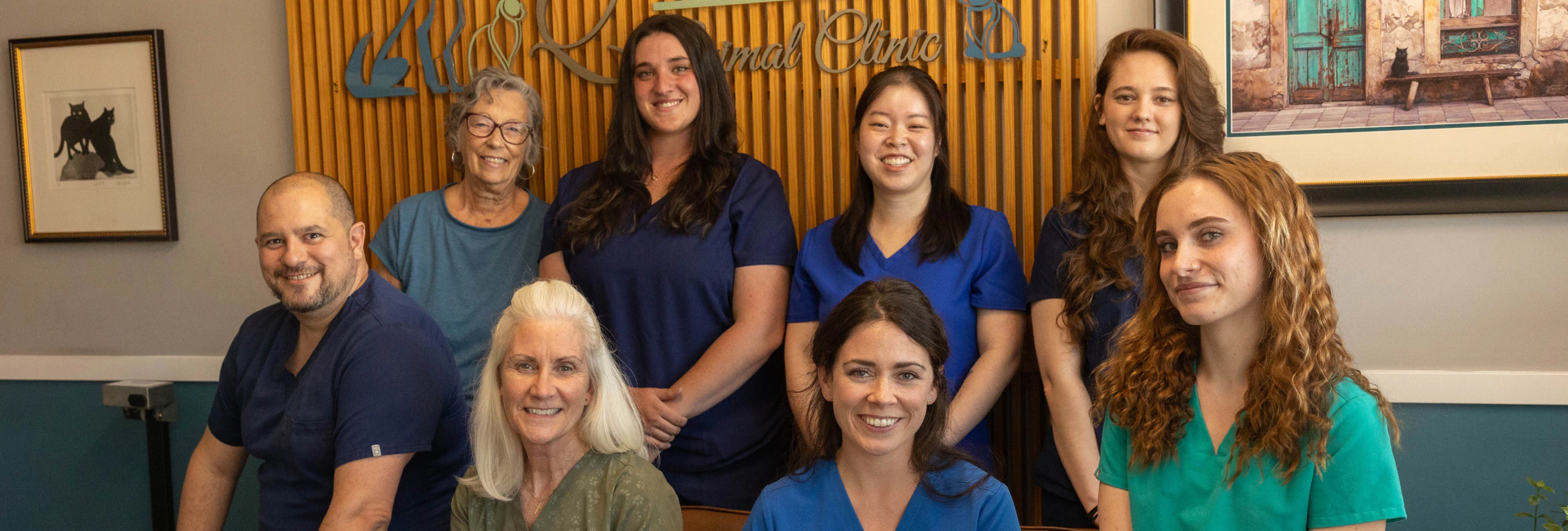 Desktop_Home_Above_Testimonials A group of eight people, some seated and some standing, smiling at the camera in a clinic setting. They wear scrubs and casual attire. There is art and a clinic sign on the wall behind them.