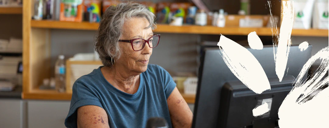 Desktop_Home_Having_A_Pet An older woman with glasses and short gray hair sits at a desk, focused on a computer screen. Shelves with various items are visible in the background. White paint-like marks appear on the right side of the image.